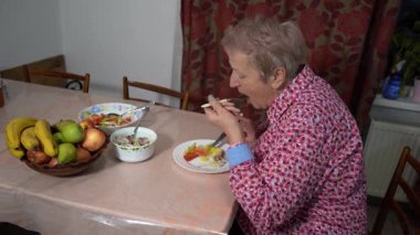 Emekli yaşlı kadın akşam yemeğini tabakta yiyor. Sağlıklı yiyecekler. Balık ve sebze. Çatal ve bıçaklı masa ve eller. Meyve kasesi.