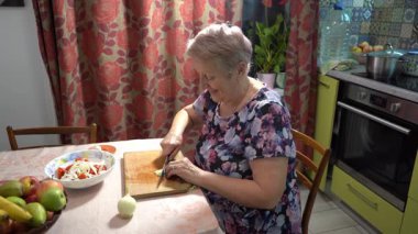 Yaşlı bir kadın mutfakta yemek hazırlıyor. Tahtada soğan kes. Sebze ve meyve salatası kesiyor..