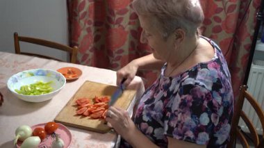 Yaşlı bir kadın mutfakta yemek hazırlıyor. Masada sebze ve meyve domates salatası kesiyor.