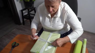 Yaşlı bir kadın hediyesini yeşil kağıda sarar. Bir büyükanne evde bir kutu toplar. Eller yukarı. Noel hazırlığı