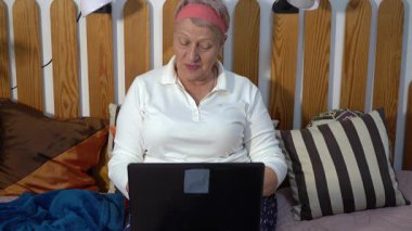 Dizüstü bilgisayarı olan aktif, yaşlı bir kadın evde yatağında otururken iletişim kuruyor ve çalışıyor. Bir büyükanne konuşur ve ekrana bakar.