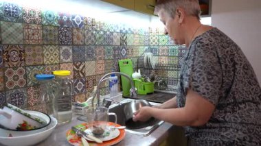 Yaşlı bir kadın mutfak lavabosunun musluğunun altında tencere yıkıyor. Kirli bulaşıkları ellerimle yıkıyorum.