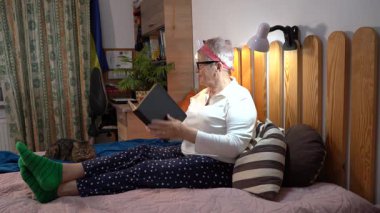 Yaşlı bir kadın yatakta oturmuş posta pullarından oluşan bir albüme bakıyor. Büyükannem ve kedisi. Evde hobi. Not defterine kalemle yaz.