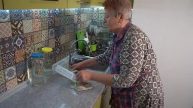 Yaşlı kadın musluğu yıkıyor ve mutfakta lavabo açıyor. Evde önlüklü bir büyükanne. Ev işi.