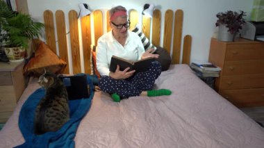Yaşlı bir kadın yatağına oturur ve kedisine sesli bir kitap okur..