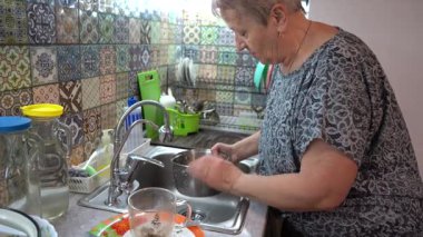 Yaşlı bir kadın mutfak lavabosunun musluğunun altında tencere yıkıyor. Kirli bulaşıkları elle yıkıyorum.