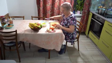 Domates doğra. Yaşlı bir kadın mutfakta yemek pişiriyor. Masada sebze ve meyve kasesi kesiyor.