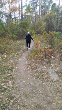 Büyükannem eşofmanla orman yolunda yürüyor. İskandinav yürüyüş ve sırıklar. Doğada yaşlı bir kadının spor aktivitesi. Geri