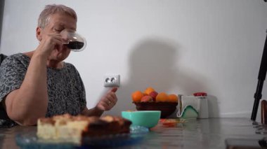 Yaşlı kadın şeffaf fincandan kahve içiyor. Büyükannem kahvaltı yapıyor. Mutfaktaki masada turta. Evde yemek var. Kahvaltıda içki.