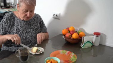 Süzme peynirli güveç turtası ye. Yaşlı kadın şeffaf fincandan kahve içiyor. Büyükannem kahvaltı yapıyor. Mutfaktaki masada meyve kasesi var. Evde yemek var..