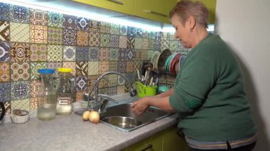 Yaşlı bir kadın patates soyar. Mutfak lavabosunda patates püresi. Evde sebze pişir. Yaşlı büyükanne iş sırasında konuşuyor.