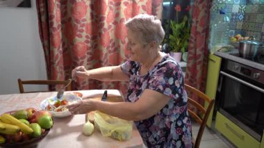 Yaşlı bir kadın salatayı kaseye koyar. Evde yemek pişirmek. Meyveler masada. Büyükanne aşçı.