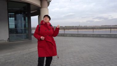 Yaşlı kadın tur rehberi nehrin yanındaki caddede duruyor ve bir hikaye anlatıyor. Kırmızı ceketli bir büyükanne konuşuyor. Açık hava.