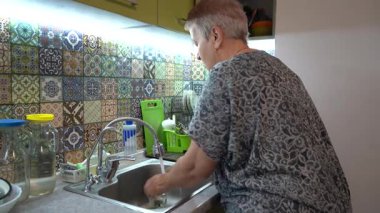 Yaşlı bir kadın mutfak lavabosundaki musluğun altında bardak ve ellerini yıkıyor. Kirli bulaşıkları ellerimle yıkıyorum..