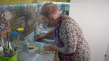 Yaşlı kadın musluğu yıkıyor ve mutfakta lavabo açıyor. Evde önlüklü bir büyükanne. Ev işi.