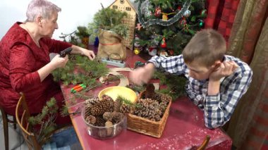 Yapıştırıcı tabancasıyla dallar ve koniler. Büyükannem ve torunum birlikte Noel çelengi yapıyorlar. Yaşlı kadın ve genç çocuk kendi elleriyle dekore ederler. Zanaat