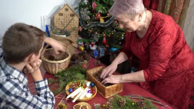 Büyükannem ve torunum birlikte Noel çelengi yapıyorlar. Yaşlı kadın ve genç çocuk kendi elleriyle dekore ederler. Aile eğlencesi. Zanaat. Kedi çalınmış koni