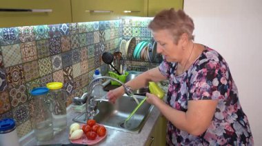 Yaşlı bir kadın mutfak lavabosunda su altında salata için sebze yıkıyor. Tatlı patates ve domates. Büyükanne yemek hazırlıyor. Evde yemek pişirmek.