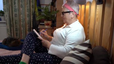 Yaşlı bir kadın yatakta oturur ve bir çizim defterine kalemle resim çizer. Bir büyükanne deftere yazar. Büyükanne ve hobisi evde.