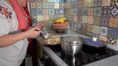 Hamur köftelerini pişir, Varenyky 'yi tavadan çıkar. Ev mutfağında yemek pişiren yaşlı bir kadın. Sobanın üstünde kaynar su buharı tenceresi. Elinde kaşık