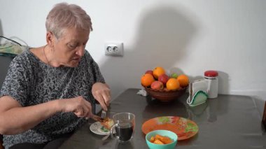 Süzme peynirli güveç turtası ye. Yaşlı kadın şeffaf fincandan kahve içiyor. Büyükanne kahvaltı yapıyor. Mutfaktaki masada meyve var. Evde yemek var. Çatal bıçak ellerinde.