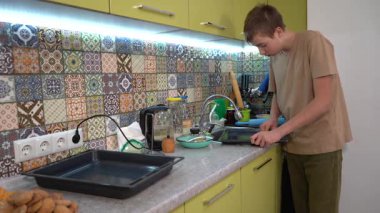 Oğlan pişirdikten sonra pişirme tepsisini yıkar. Ergen, mutfak lavabosunda bulaşıkları yıkıyor. Ev işleri.