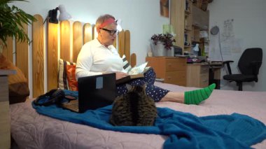 Yaşlı bir kadın yatakta oturmuş posta pullarından oluşan bir albüme bakıyor. Büyükanne ve kedi hobisi evde.