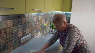 Yaşlı kadın musluğu yıkıyor ve mutfakta lavabo açıyor. Evde önlüklü bir büyükanne. Ev işi.