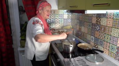 Yaşlı kadın çeşitli hamur işleri pişiriyor. Ukrayna yemeği. Nakışlı gömlekli büyükanne. Ocakta bir tencere kaynıyor. Mutfak evde..