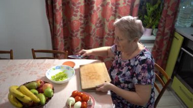 Yaşlı bir kadın mutfakta yemek pişiriyor. O, sebze ve meyve salatası kesiyor. Yeşil kağıt ve domates kasesi