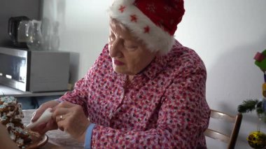 Kırmızı Noel Baba şapkalı yaşlı kadın zencefilli kurabiye ağacını beyaz kremayla süslüyor. Ev kurabiyelerinde yemek pişirmek. Noel pastası. Kış yemeği. Büyükanne yemek yapıyor.