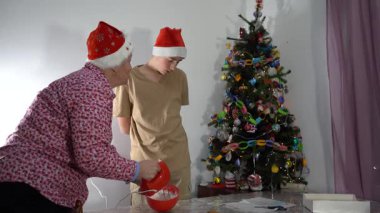 Kırmızı Noel Baba şapkalı yaşlı kadın karıştırıcı ile kurabiye için krema yapıyor. Büyükannenin evinde Noel ağacı. Bir çocuk büyükannesiyle evde kurabiye pişirir. Yiyecek.