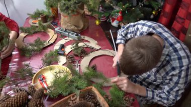 Noel çelengi yap. Genç çocuk zamk dalları kullanıyor. Kışlık ev yapımı masa manzarası. Noel ağacına yakın bir aile. DIY hobisi