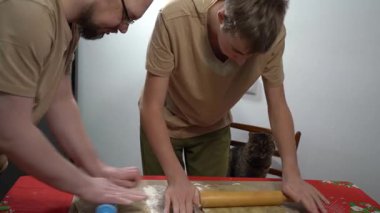 Hamuru merdaneyle fırlat. Erkek ve genç bir çocuk hamurdan kurabiye yapıyorlar. Baba ve oğul birlikte yemek pişiriyorlar. Baba aşçı. Evde kedi var..
