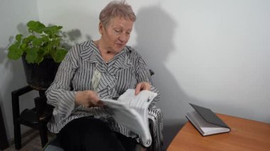 Yaşlı bir kadın bir sandalyede oturuyor ve ders kitabı okuyor. Bir büyükanne kitap okuyor. Ders kitabında yazıyor. Evde çalış.