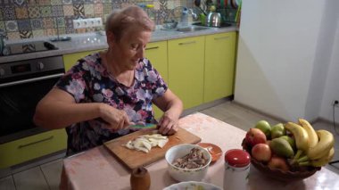 Tahtada soğan doğra. Yaşlı bir kadın mutfakta yemek hazırlıyor. Sebze ve meyve salatası kesiyor..