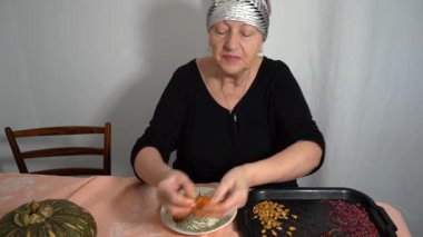 Kurutmak için kabak çekirdeği topluyorum. Yaşlı bir kadın böğürtlen tohumlarını kurutuyor. Bir büyükanne mutfak masasında oturur ve yemek pişirir. Kuru yiyecek.