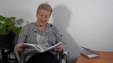 Yaşlı bir kadın bir sandalyede oturuyor ve ders kitabı okuyor. Bir büyükanne kitap okuyor ve gülüyor. Evde çalış.