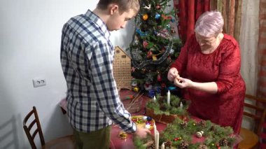 Büyükannem ve torunum birlikte Noel çelengi yapıyorlar. Yaşlı bir kadın ve genç bir çocuk. Koniler, ladin dalları ve mumlar. Yılbaşı ağacı evde. Aile kış zamanı.