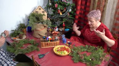 Büyükannem Noel çelengi yapar. Yaşlı kadın işi. Evdeki köknar dallarını Noel ağacı toplarıyla süsleyin. Kış aktiviteleri. Çelenk dekorasyonu