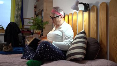 Yaşlı bir kadın kedisiyle yatakta oturuyor, posta pullarından oluşan bir albüme bakıyor. Büyükanne ve hobisi evde.