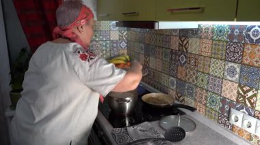 Hamur köftesi pişiren yaşlı bir kadın. Ukrayna yemeği. Nakışlı gömlekli bir büyükanne. Ocakta bir tencere kaynıyor. Evde bir mutfak..