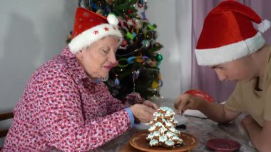 Kırmızı Noel Baba şapkalı yaşlı kadın zencefilli kek ağacını kremayla süslüyor. Ergen torunumla evde yemek pişirmek. Noel pastası. Kış yemeği.