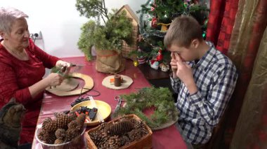 Büyükannem ve torunum birlikte Noel çelengi yapıyorlar. Yaşlı kadın ve genç çocuk kendi elleriyle dekore ederler. Aile eğlencesi. Zanaat. Çam dalları