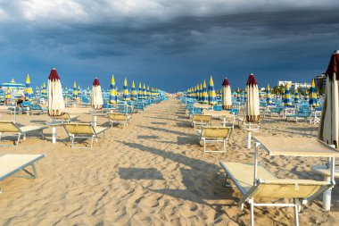 Kumsalda, Şemsiyeli ve Güverte Sandalyeli. Yaz tatili. Fırtınadan önce gökyüzü. Boş sahil şeridi şezlongu. Sarı mavi şemsiye güneş banyosu.