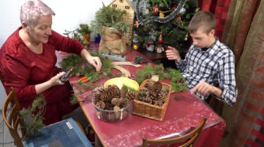 Büyükannem ve torunum birlikte Noel çelengi yapıyorlar. Yaşlı kadın ve genç çocuk dekore ediyor. Süslü dallar ve koniler. Aile Zirvesi.