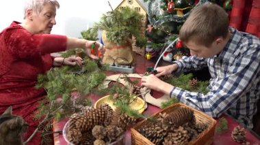 Büyükannem ve torunum birlikte Noel çelengi yapıyorlar. Yaşlı kadın ve genç çocuk çam dallarını yapıştırıyor. Aile eğlencesi. Zanaat