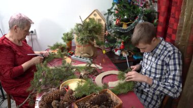 Büyükannem ve torunum birlikte Noel çelengi yapıyorlar. Yaşlı kadın ve genç çocuk Noel ağacı dallarını ve konilerini yapıştırıyor. Aile içi kış aktiviteleri..