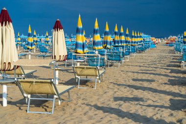 Şemsiyelerle kumlu sahilde güneşleniyorlar. Boş sahil beldesi. İtalya. Sahil şeridi sıra şezlongda güneşleniyor. Yazın mavi yağmurlu gökyüzü