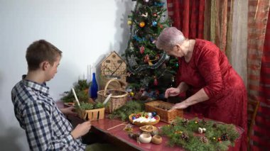 Büyükannem ve torunum birlikte Noel süsü yapıyorlar. Yaşlı kadın ve genç çocuk Noel ağacının yanında. Mumlar ve çam dalları Aile kış gemisi